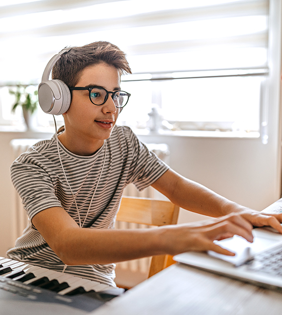 Boy wearing headphones sitting in front of piano at home with laptop for online lessons