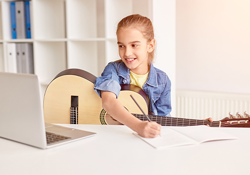 A girl sitting with a guitar at a desk with a laptop making notes