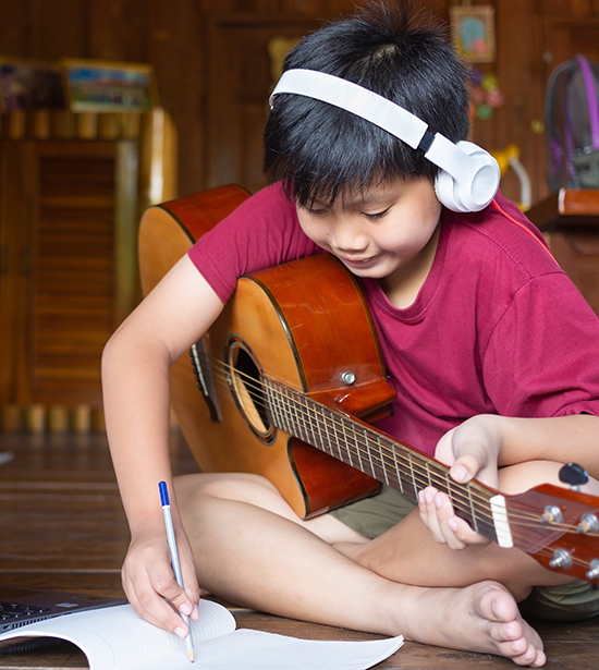 A boy sitting with a guitar wearing headphones by a laptop making notes