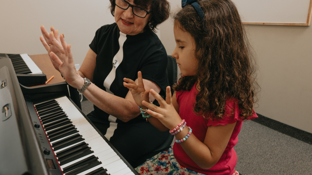A young girl smiling during a piano lesson at The Piano Studio, seated at a piano with a teacher beside her.