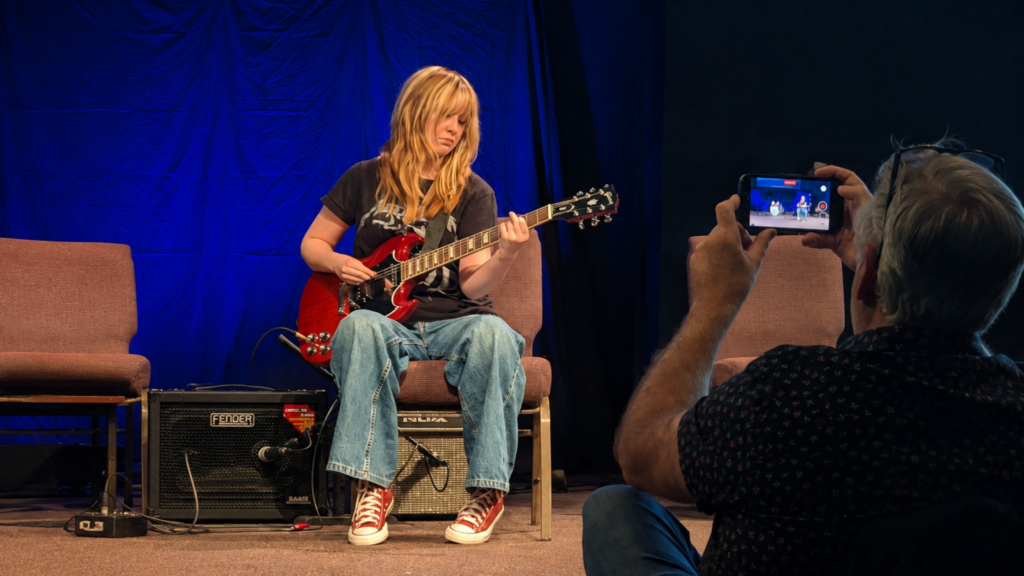 A young guitar student plays electric guitar on stage while a parent records the moment on their phone.